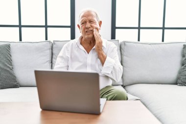 Evde dizüstü bilgisayar kullanan yaşlı bir adam diş ağrısı ya da diş hastalığından dolayı kanepede oturup ağızlarına acı dolu bir ifadeyle dokunuyor. dişçi 