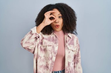 Young african american woman standing over blue background doing ok gesture shocked with surprised face, eye looking through fingers. unbelieving expression. 