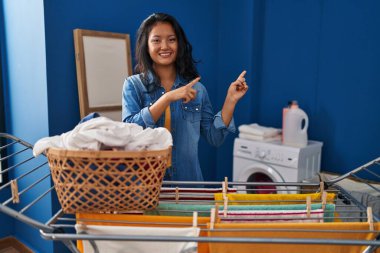 Young asian woman hanging clothes at clothesline smiling and looking at the camera pointing with two hands and fingers to the side. 