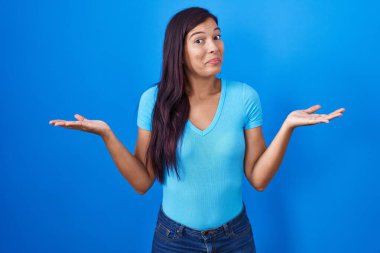 Mavi arka planda dikilen Latin kökenli genç kadın elleri ve kolları havada, kafası karışmış bir ifade takınıyor. şüphe kavramı. 