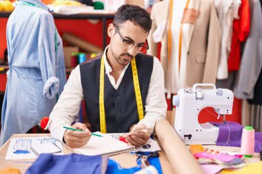 Young hispanic man tailor holding cloths drawing on notebook at atelier