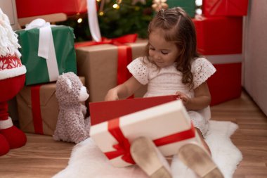 Adorable hispanic girl unpacking gift sitting on floor by christmas tree at home