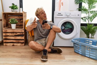 Young blond man doing laundry using smartphone pointing fingers to camera with happy and funny face. good energy and vibes. 