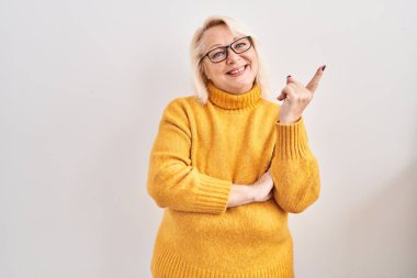 Middle age caucasian woman wearing glasses standing over background with a big smile on face, pointing with hand and finger to the side looking at the camera. 