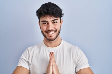 Beyaz arka planda sakallı İspanyol bir adam elleriyle dua edip af diliyor kendinden emin bir şekilde gülümseyerek.. 