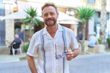 Middle age man smiling confident holding sunglasses at coffee shop terrace