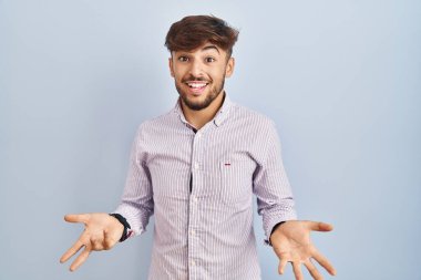 Arab man with beard standing over blue background smiling cheerful with open arms as friendly welcome, positive and confident greetings 