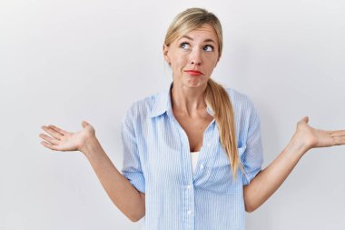 Beautiful blonde woman standing over white background clueless and confused expression with arms and hands raised. doubt concept. 