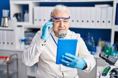 Middle age grey-haired man scientist talking on smartphone using touchpad at laboratory