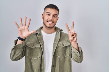 İzole edilmiş arka planda duran genç İspanyol adam yedinci parmaklarını gösterip işaret ederken kendinden emin ve mutlu gülümsüyor.. 