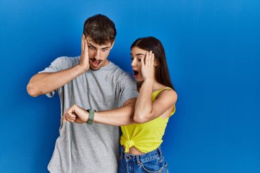 Young hispanic couple standing together over blue background looking at the watch time worried, afraid of getting late 