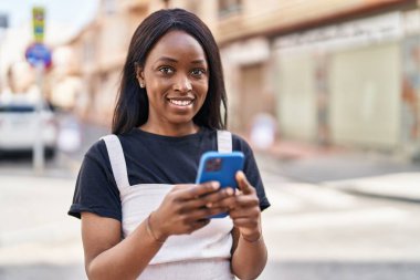 Genç Afro-Amerikalı kadın sokakta akıllı telefon kullanarak gülümsüyor.