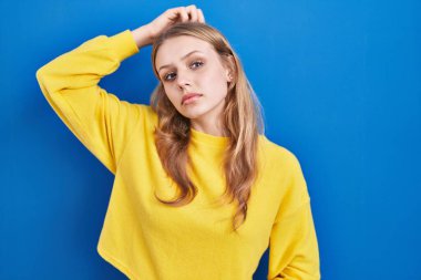 Young caucasian woman standing over blue background confuse and wondering about question. uncertain with doubt, thinking with hand on head. pensive concept. 