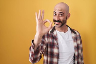 Sarı arka planda duran sakallı İspanyol adam gülümsüyor ve el ve parmaklarıyla 