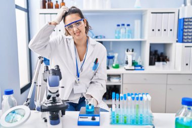 Young hispanic woman working at scientist laboratory confuse and wonder about question. uncertain with doubt, thinking with hand on head. pensive concept. 