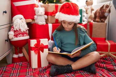 Adorable hispanic girl writing on notebook sitting on floor by christmas tree at home