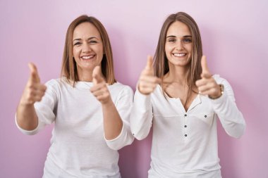 Middle age mother and young daughter standing over pink background pointing fingers to camera with happy and funny face. good energy and vibes. 