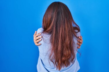Hispanic young woman standing over blue background hugging oneself happy and positive from backwards. self love and self care 