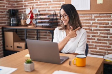 Hispanic young woman working at the office wearing glasses smiling cheerful pointing with hand and finger up to the side 