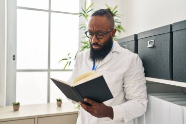 Young african american man wearing doctor uniform reading book at clinic
