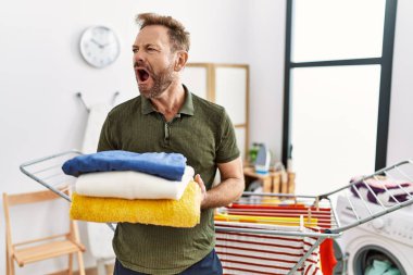 Middle age man holding folded laundry after ironing angry and mad screaming frustrated and furious, shouting with anger. rage and aggressive concept. 