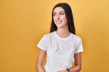 Young beautiful woman standing over yellow background looking away to side with smile on face, natural expression. laughing confident. 
