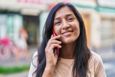 Genç, güzel, İspanyol bir kadın gülümsüyor ve caddede akıllı telefondan konuşuyor.