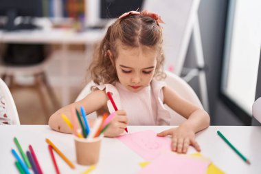 Sevimli beyaz kız öğrenci sınıftaki masanın üzerine oturmuş, kağıda resim çiziyor.