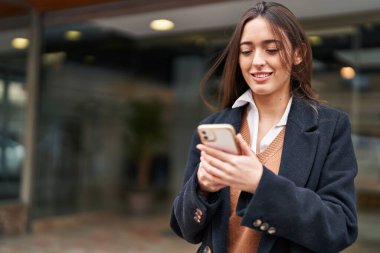 Genç, güzel, İspanyol bir kadın sokakta akıllı telefon kullanarak gülümsüyor.