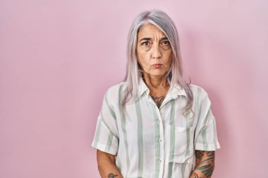 Pembe arka planda duran gri saçlı, kuşkucu ve gergin orta yaşlı bir kadın. Yüzünde çapraz kollu onaylamayan bir ifade var. negatif kişi. 