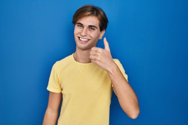 Young man standing over blue background doing happy thumbs up gesture with hand. approving expression looking at the camera showing success. 
