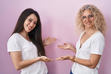 Pembe arka planda yan yana duran anne ve kızı gülümseyerek doğal bir şekilde içeri girmeye davet ediyorlar. 