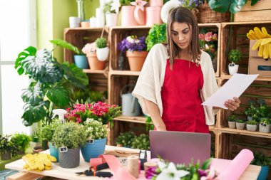 Young beautiful hispanic woman florist using laptop reading document at flower shop