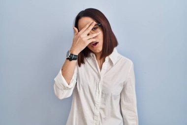 Young hispanic woman standing over white background peeking in shock covering face and eyes with hand, looking through fingers with embarrassed expression. 