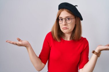 Young redhead woman standing wearing glasses and beret clueless and confused expression with arms and hands raised. doubt concept. 