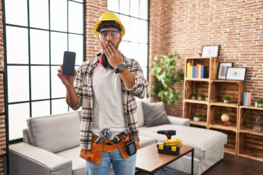 Evde tadilat yapan sakallı genç İspanyol bir adam elinde akıllı bir telefonla ağzını eliyle kapatıyor, şok geçiriyor ve hata yapmaktan korkuyor. şaşırmış ifade 