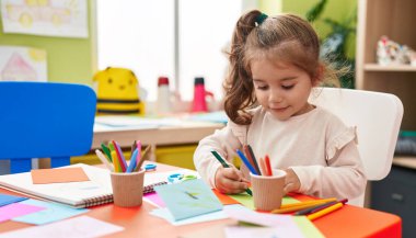 Anaokulunda masaya oturmuş kağıda resim çizen sevimli İspanyol kız öğrenci.