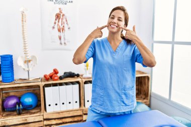 Middle age hispanic physiotherapist woman working at pain recovery clinic smiling cheerful showing and pointing with fingers teeth and mouth. dental health concept. 