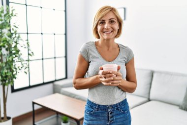 Orta yaşlı, sarışın, kendine güvenen, evde kahve içen bir kadın.