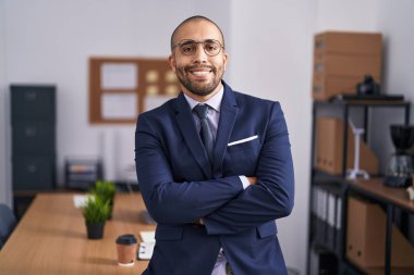 Young latin man business worker standing with arms crossed gesture at office