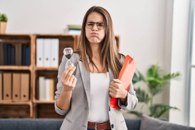 Beautiful woman working at therapy office holding sand clock puffing cheeks with funny face. mouth inflated with air, catching air. 
