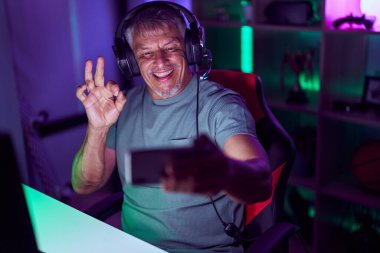 Hispanic man with grey hair playing video games with smartphone doing ok sign with fingers, smiling friendly gesturing excellent symbol 