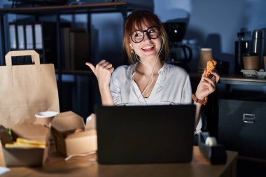 Young beautiful woman working using computer laptop and eating delivery food smiling with happy face looking and pointing to the side with thumb up. 