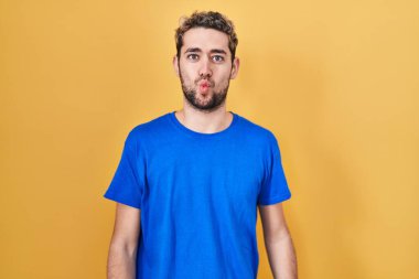 Hispanic man with beard standing over yellow background making fish face with lips, crazy and comical gesture. funny expression. 