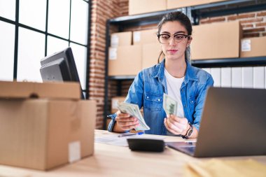 Young beautiful hispanic woman ecommerce business worker counting dollars using laptop at office