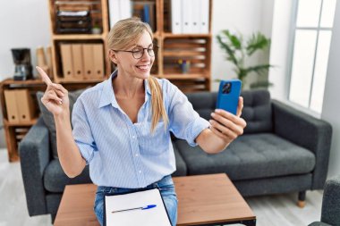 Beautiful woman doing online therapy session at consultation office smiling happy pointing with hand and finger to the side 