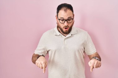 Plus size hispanic man with beard standing over pink background pointing down with fingers showing advertisement, surprised face and open mouth 