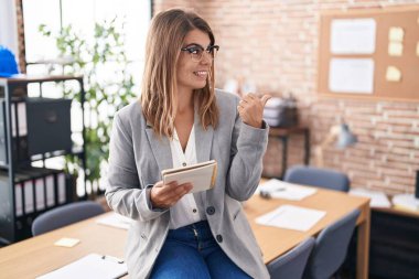 Ofiste çalışan genç İspanyol kadın. Gözlüklü, mutlu yüzlü, gülümseyen ve başparmağıyla yan tarafı gösteren.. 