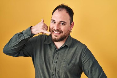 Plus size hispanic man with beard standing over yellow background smiling doing phone gesture with hand and fingers like talking on the telephone. communicating concepts. 