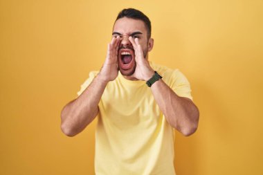 Sarı arka planda dikilen Latin asıllı genç adam elleri ağzında kızgın bir şekilde bağırıyor. 